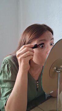 Woman applies mascara while looking in a mirror during daytime in a