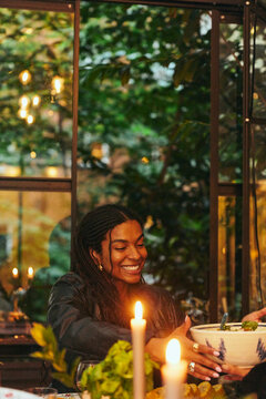Smiling young woman passing bowl of food at illuminated dinner table in courtyard