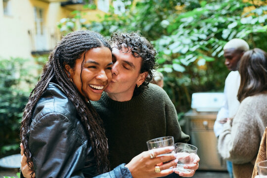 Affectionate young man kissing girlfriend on cheek in courtyard party
