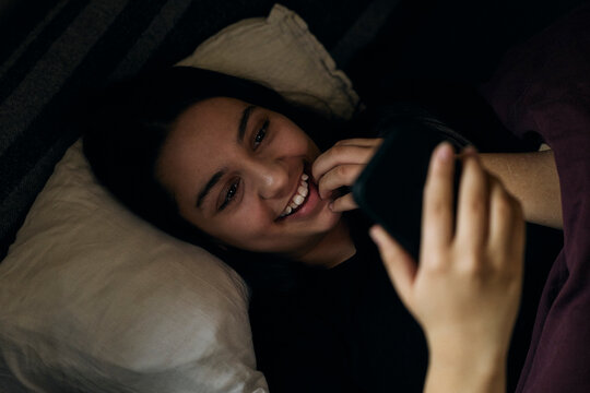 High angle view of happy girl using social media on her smart phone while lying in bed