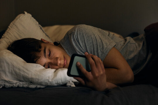 Teenage boy scrolling smart phone while lying in bed before sleeping
