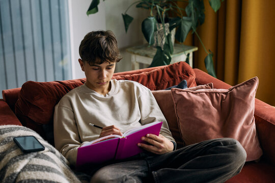 Teenage boy writing in planner while sitting on couch at home
