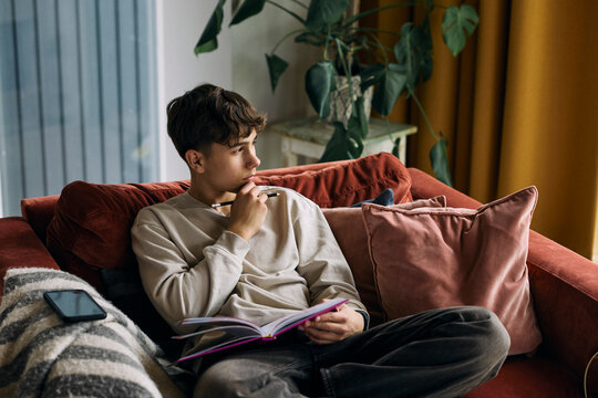 Thoughtful teenage boy with hand on chin sitting with planner on sofa at home