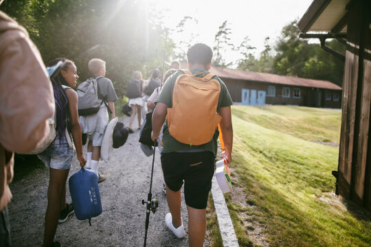 Rear view of walking on dirt path while carrying backpacks and gear at summer camp