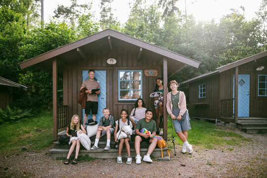 Portrait of happy children and counselors posing together on porch of wooden cabin at summer camp