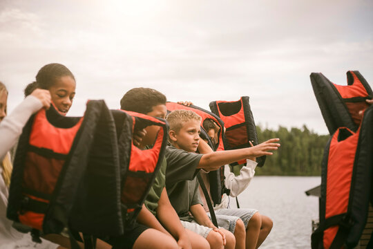 Group of children putting on red and black life jackets while sitting by lake