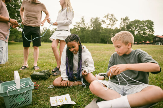 Boy and girl practicing tying knots with ropes while sitting on grass at summer camp