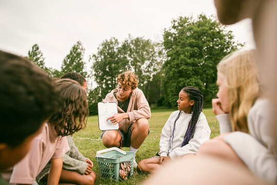 Adult female instructor pointing to drawing of compass on small clipboard while teaching kids at summer camp