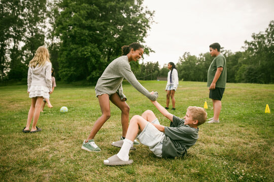 Girl lending helping hand to boy lying on grass during fun activity at park