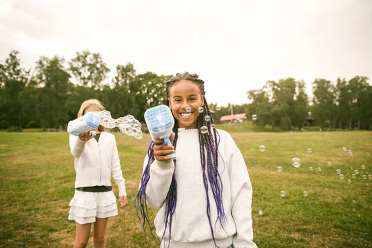 Diverse happy girls playing with bubble blower toy on grassy field at park