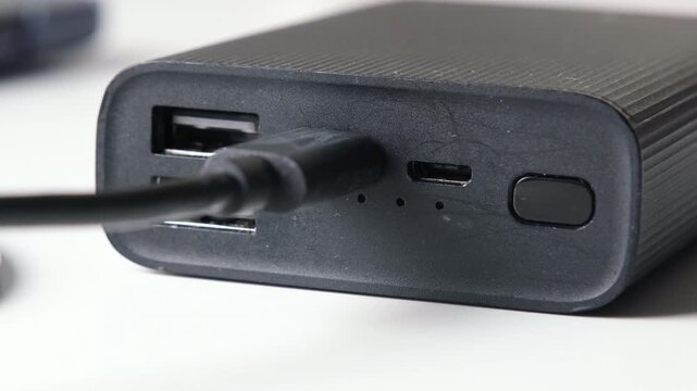 Close-up of a black portable power bank charging through a connected cable, showing usb ports, battery indicator lights and power button on a white background