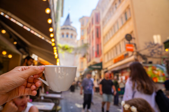 Hand holding a white coffee cup in a bustling street with colorful buildings and the Galata Tower visible in the background