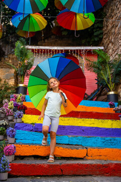 Young girl with a colorful umbrella walks down vibrant steps adorned with flowers and hanging umbrellas in a lively outdoor setting