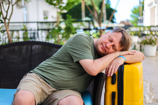 Tourist sleeping on suitcase in hotel lobby waiting airport transfer, tired traveler rest, vacation end, luggage, journey concept.