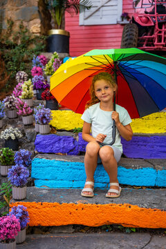 Young girl with rainbow umbrella sits on colorful steps surrounded by vibrant flowers in a lively outdoor setting