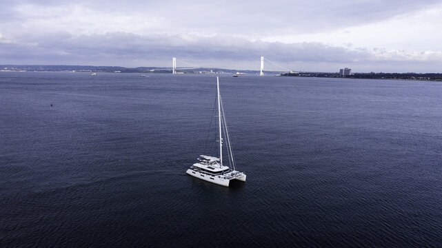 Aerial view of a white catamaran sailing on the calm blue waters of the bay with the Verrazzano-Narrows Bridge in the background in Kings County, New York, United States.