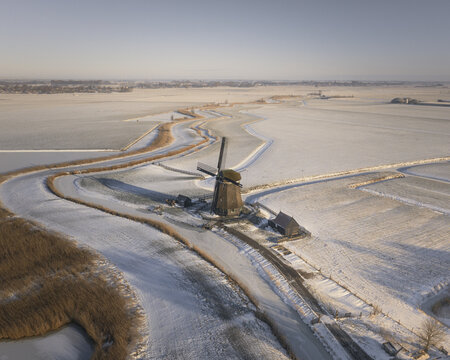 Aerial view of a traditional Dutch windmill standing beside a winding frozen canal in a snow-covered landscape in Ursem, North Holland, Netherlands.