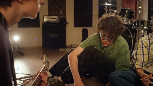 Caucasian teenage musicians sitting on floor discussing sheet music then fist bumping during rock band rehearsal. Clip for youth musical education, teen creativity, or music school marketing campaigns