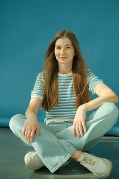 Young happy blonde woman with long healthy hair in striped top and jeans sitting on the floor in studio on blue background