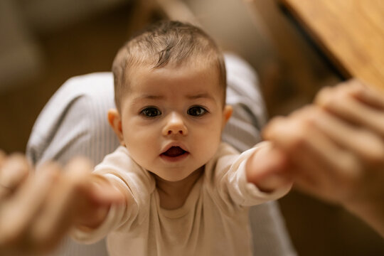 Baby reaches out with hands toward adult hands in warm home setting