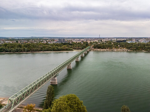 Aerial view of a green steel railway bridge crossing the Danube beside tree-lined Nepsziget, with urban neighborhoods and riverside greenery in Budapest, Hungary.