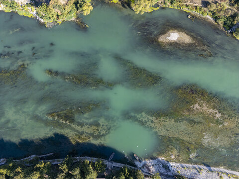 Aerial view of the Buna-Drin River Confluence showing clear turquoise waters, riverbed vegetation, and small boats moored along the tree-lined shore in Zues, Qarku i Shkodres, Albania.