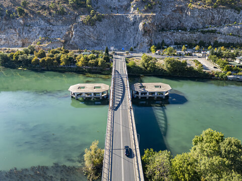 Aerial view of Ura e re e Bunes bridge spanning the Buna River with a car driving across, surrounded by lush trees and rocky hills in Shkoder, Qarku i Shkodres, Albania.