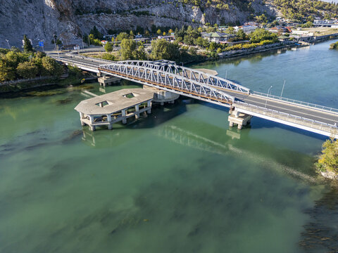 Aerial view of Ura e re e Bunes bridge spanning the turquoise Buna River, with lush greenery, rocky cliffs, and urban infrastructure in Shkoder, Qarku i Shkodres, Albania.