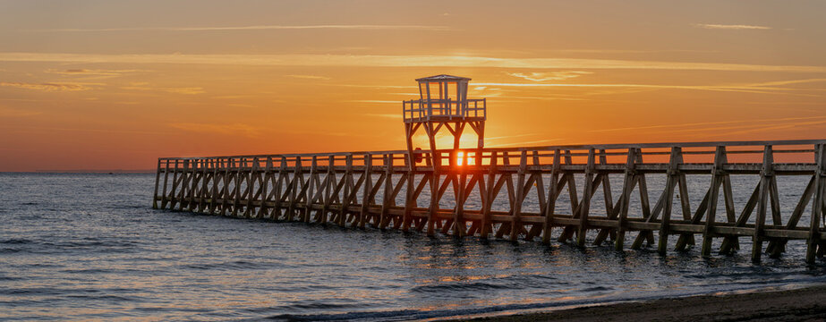 Luc-Sur-Mer, France - 08 08 2026:  Panoramic view of a wooden pier in front of the beach, sea, a colorful sunrise and Le Havre city far away