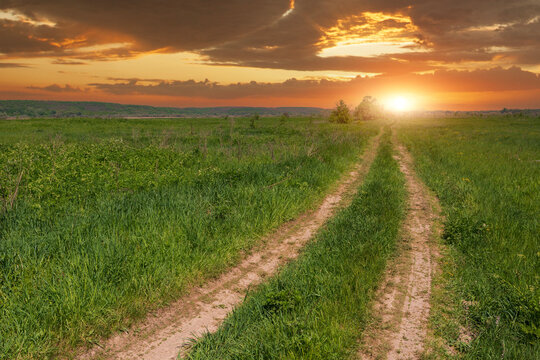 A scenic dirt path winding through a lush green meadow under a vibrant golden sunset sky