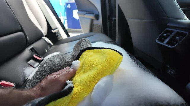 Wiping cleaning foam from car seat with microfiber cloth close-up, professional auto detailing and interior chemical cleaning service, man hand washing car upholstery with thick foam.