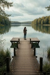 Fototapeta premium Young woman stands on wooden dock talking on smartphone. Enjoys calm lake scenery under cloudy sky. Lone female person communicates via mobile phone by quiet forest river. Peaceful nature landscape