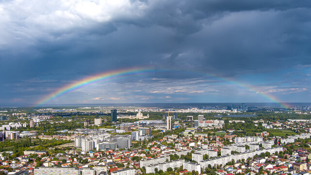 Aerial view of a vibrant rainbow arching over the cityscape of Bucharest, featuring residential blocks and green spaces under a dramatic stormy sky in Bucharest, Bucharest, Romania.