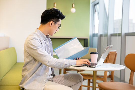 Young Asian Man Working on Laptop and Reviewing Documents in a Modern Cafe