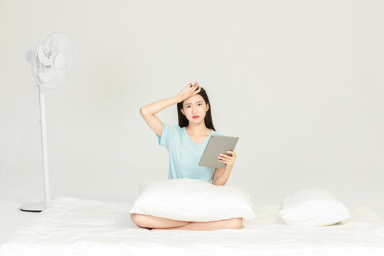 Young woman feeling hot and unwell while using a tablet on a bed near an electric fan