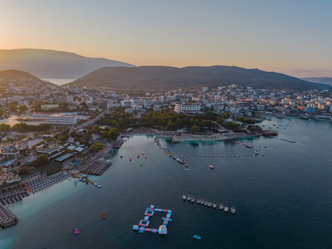 Aerial view of the coastal town of Ksamil with its beaches, floating water park, and boats in the bay, set against a backdrop of mountains in Ksamil, Vlore County, Albania.