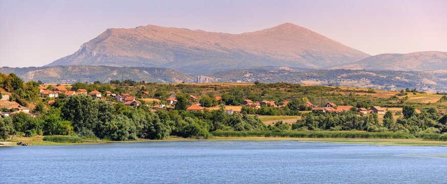 Rural village with red roofs nestled among green trees and hills, overlooking a calm lake with Rtanj mountain in the background
