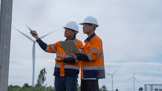 Two Asian male engineers wearing hardhats and high visibility orange jackets discussing field operations with a laptop and walkie talkie at a wind turbine power station.