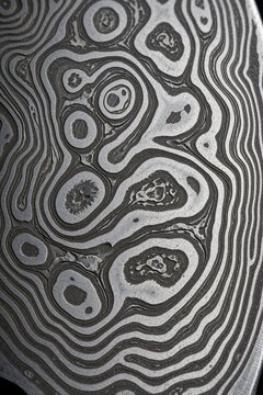 Macro shot of damascus steel blade surface. Wavy organic patterns on grey metal texture show unique forged design. Close up view of sharp knife material with silver metallic lines, dark etched