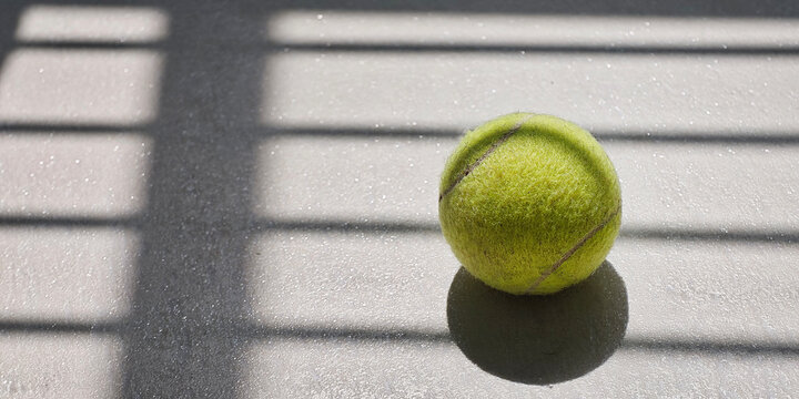 Tennis ball resting on a textured court surface, receiving morning sunlight and creating a graphic shadow pattern