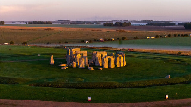 Aerial view of the ancient stone circle of Stonehenge surrounded by lush green fields and rolling hills during the golden hour in Salisbury, England, United Kingdom.