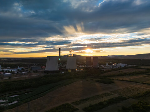 Aerial view of the Matra power plant with cooling towers and a chimney releasing steam at sunset, amid farmland and the Matra hills near Visonta, Hungary.