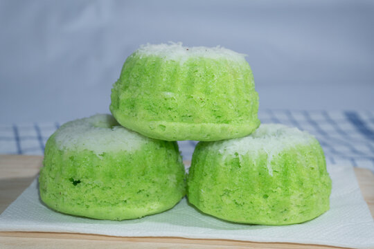 Three green putu ayu steamed cakes stacked on a wooden board, showing their soft texture and pandan-colored surface against a blue and white checkered cloth.