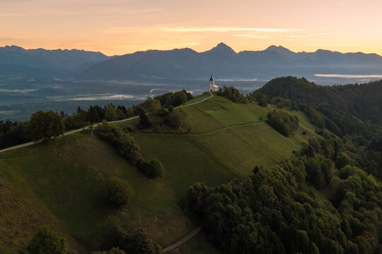 Aerial view of the Church of Saints Primus and Felician perched on a grassy hill with a winding path and mountain backdrop during golden hour in Jamnik, Kranj, Slovenia.