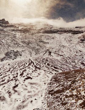Aerial view of the snow-covered slopes and rocky terrain of Volcano Chimborazo under a cloudy sky in Guano, Chimborazo, Ecuador.