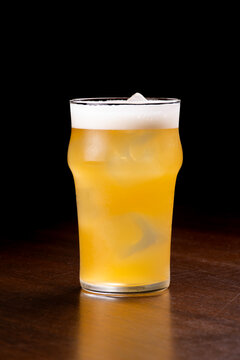 Close-up of a half-pint glass of Radler beer with ice on a wooden table, black background.