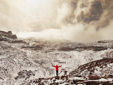 Aerial view of a person in a red jacket standing with arms outstretched on the snowy, rocky slopes of Volcano Chimborazo in Guano, Chimborazo, Ecuador.