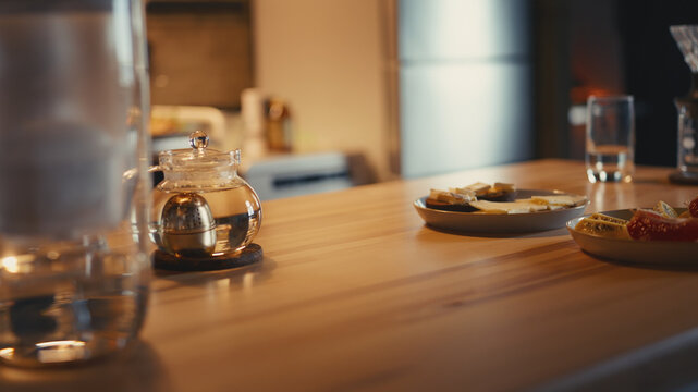 Healthy dinner on a wooden kitchen table with cheese open sandwiches, sliced fruits, and a glass teapot with brewing herbal tea. Table prepared for an upcoming healthy meal in a warm cozy home.