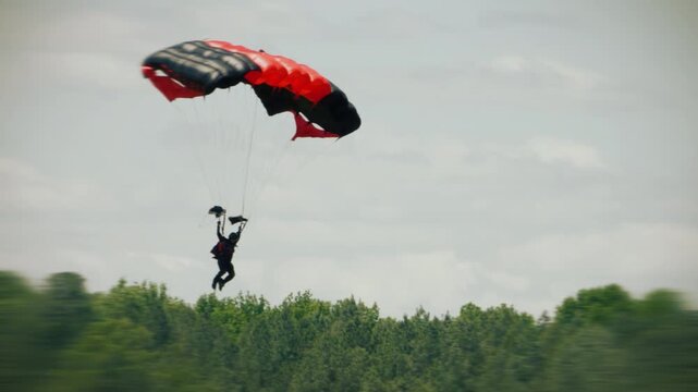 Two tandem skydivers descend beneath red and black parachute canopy in open sky, holding harness grips during coordinated freefall over cloudscape bright daylight