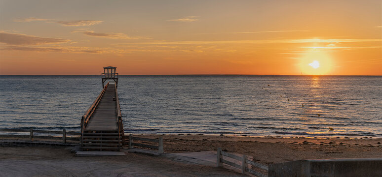 Luc-Sur-Mer, France - 08 08 2026:  View of a wooden pier in front of the sea, a colorful sunrise and port of Le Havre far away